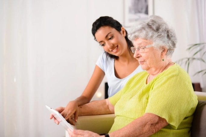 A woman is providing home care services to an older woman by sitting on a couch and looking at a tablet.