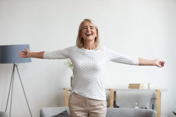 A senior woman is standing with her arms outstretched in the living room, promoting quality of life.