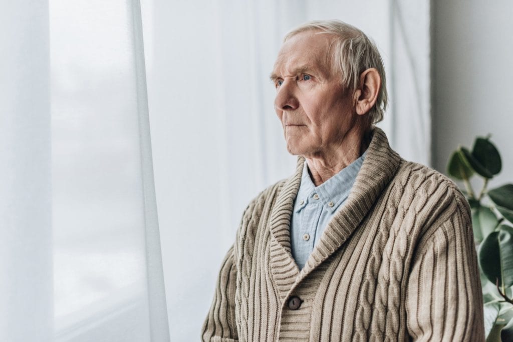 An elderly man is standing by a window.