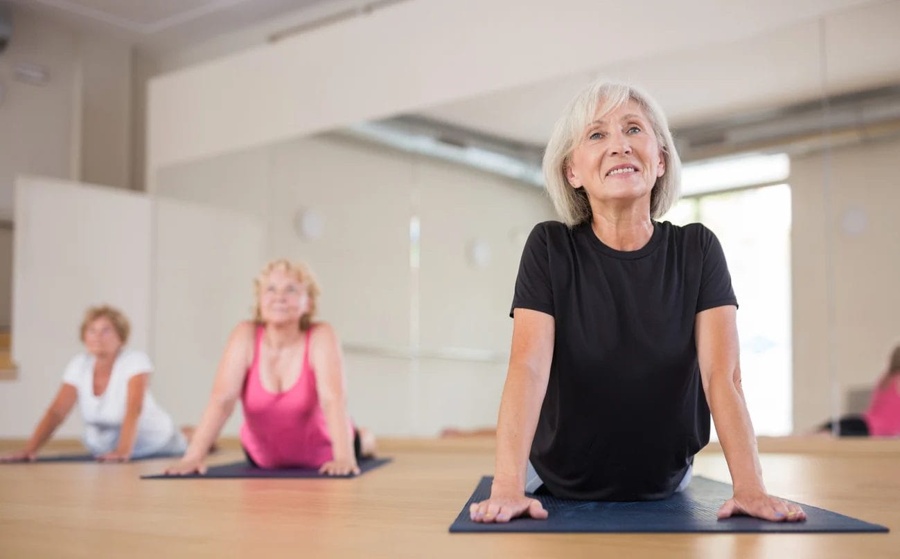 Three women are practicing yoga on mats in a bright room, performing the cobra pose. The woman in front wears a black shirt, while the two women behind her wear pink and white tops.