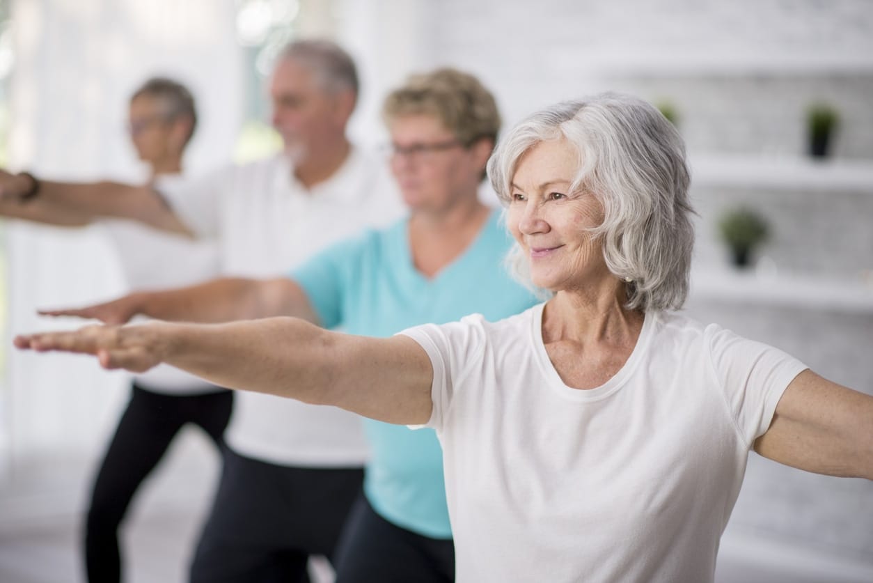 A group of four senior people performs yoga in a bright room, with a focus on a white-haired woman in the foreground extending her arms in a warrior pose.