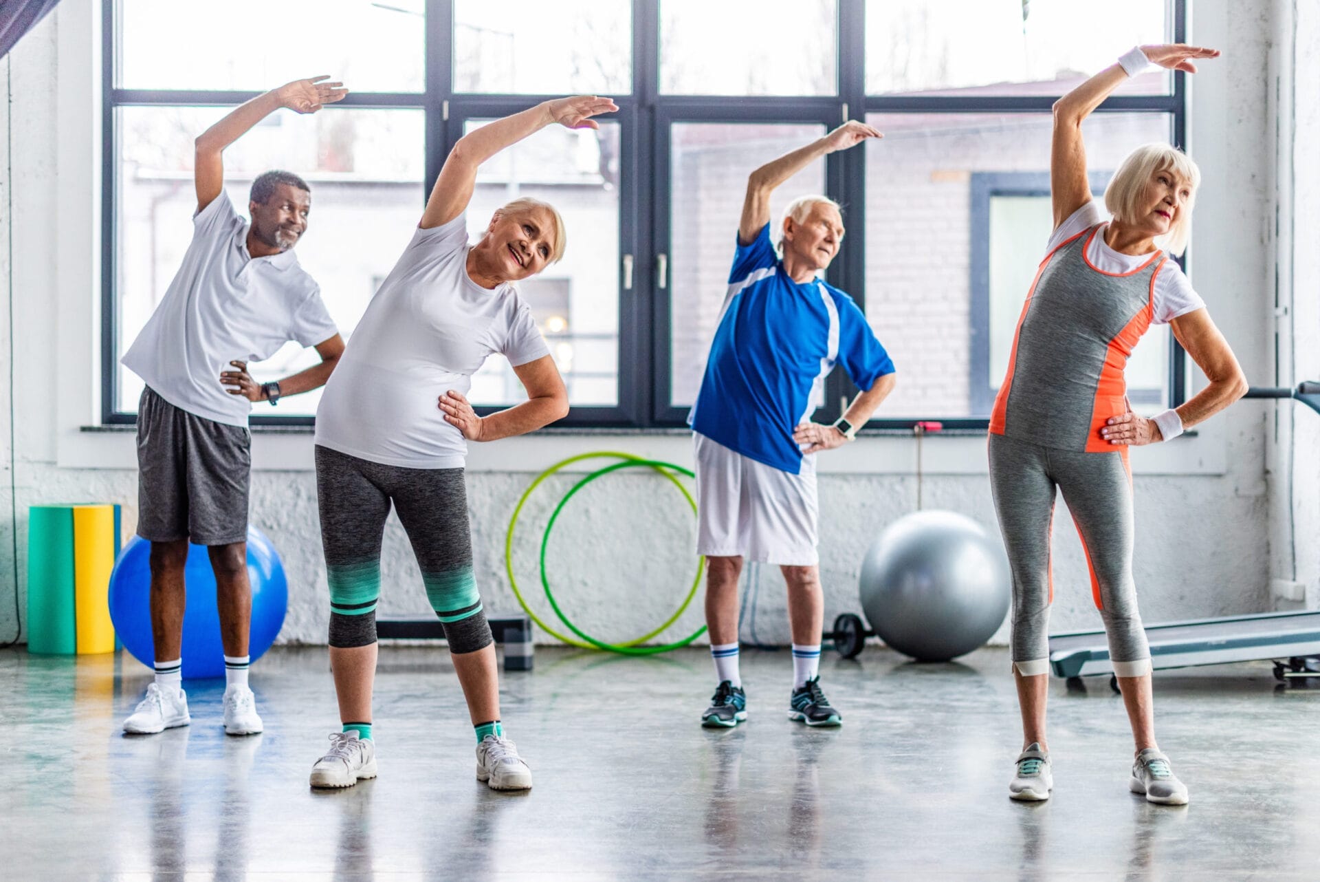 Four elderly individuals are performing side stretches in a fitness studio with large windows and various exercise equipment in the background.