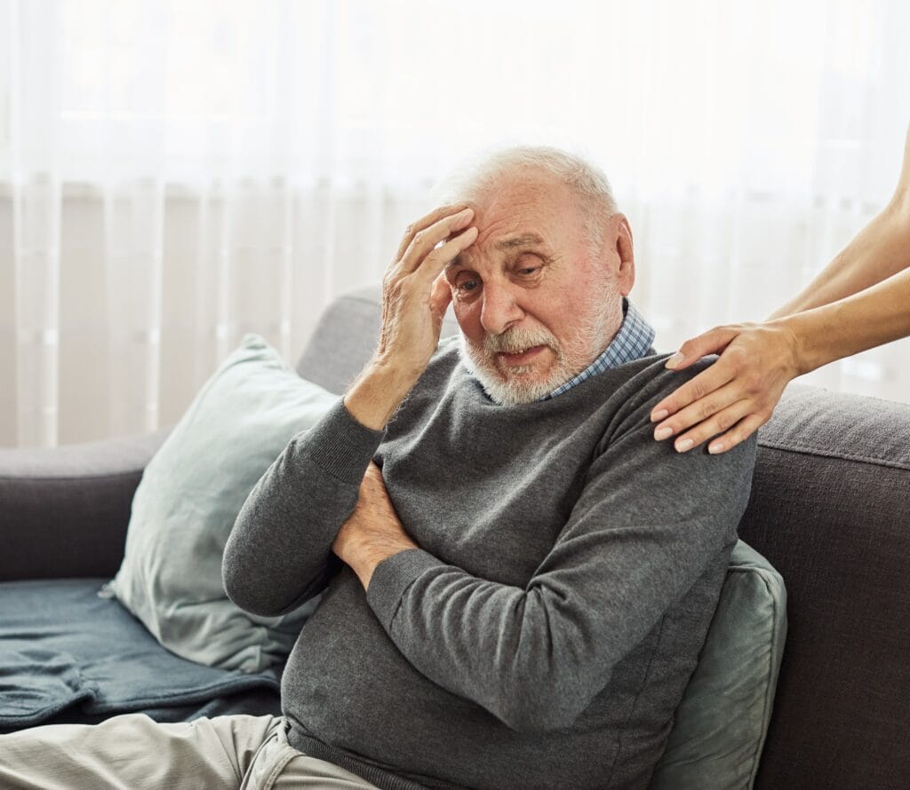 An elderly man sitting on a couch holds his head in one hand, looking distressed, while another person places a comforting hand on his shoulder.