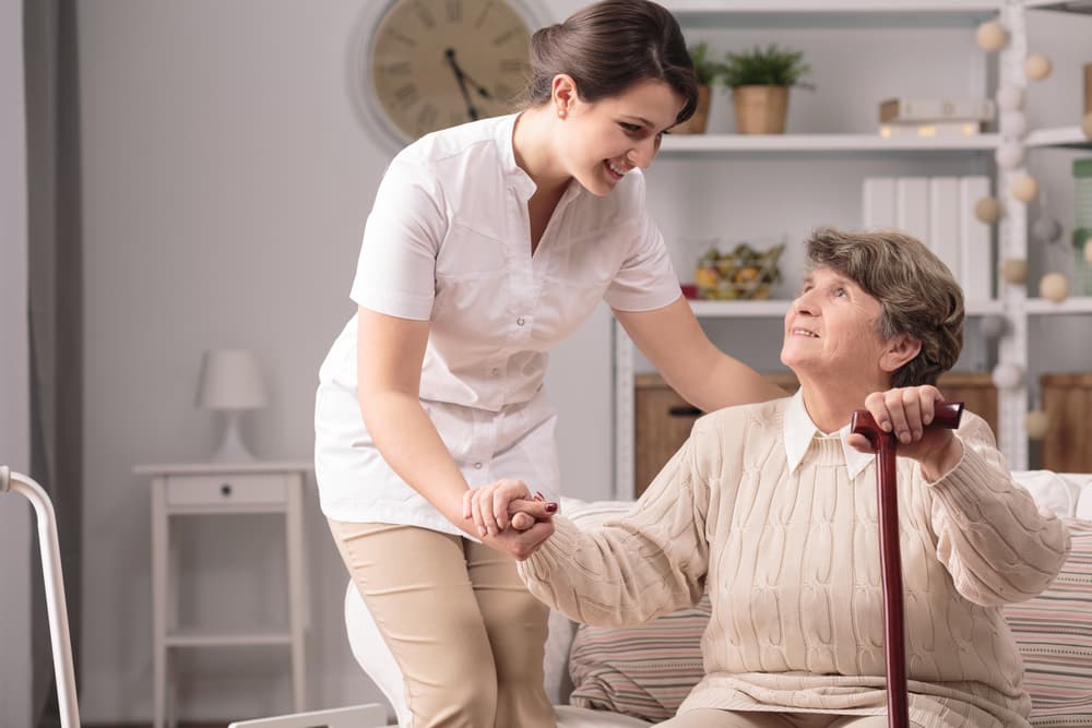 A young caregiver helps an elderly woman with a cane stand up in a bright, neatly furnished room.