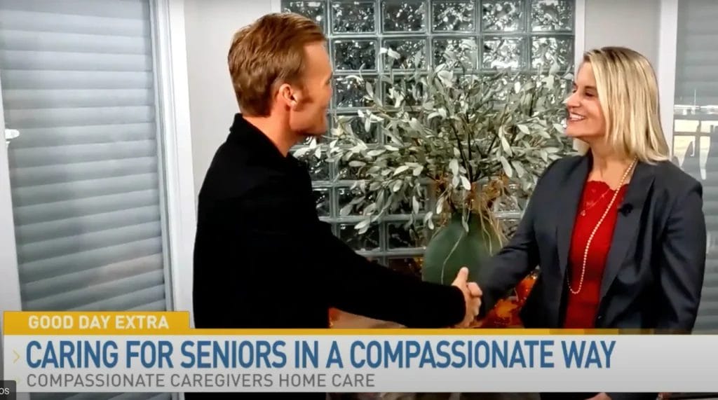 Two people shake hands and smile during a TV interview segment about compassionate home care for seniors, with a plant and glass block wall in the background.