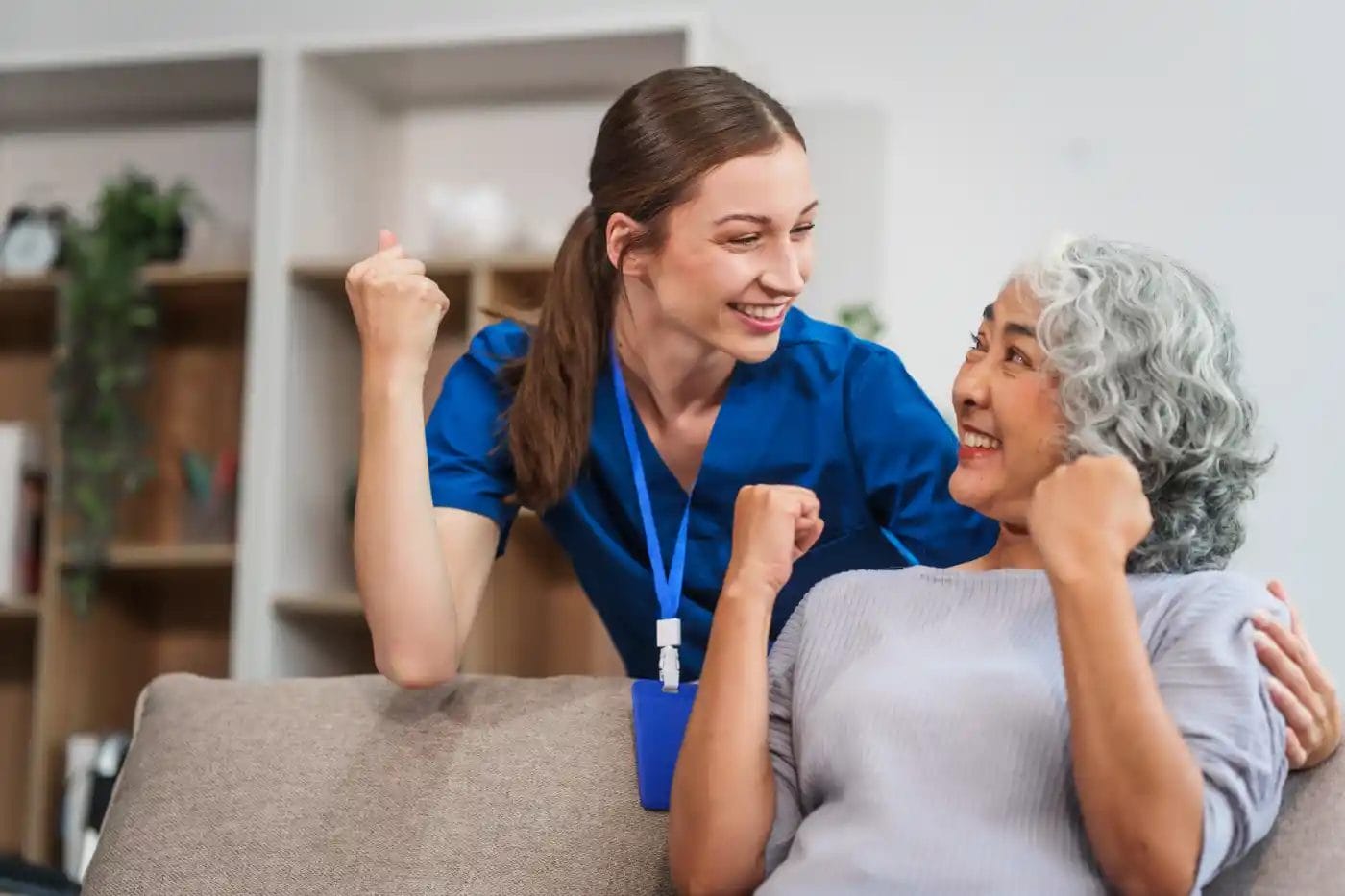 A smiling healthcare worker and an older woman sit on a couch, both raising their fists in a celebratory gesture, reflecting the positive impact of quality nursing services.