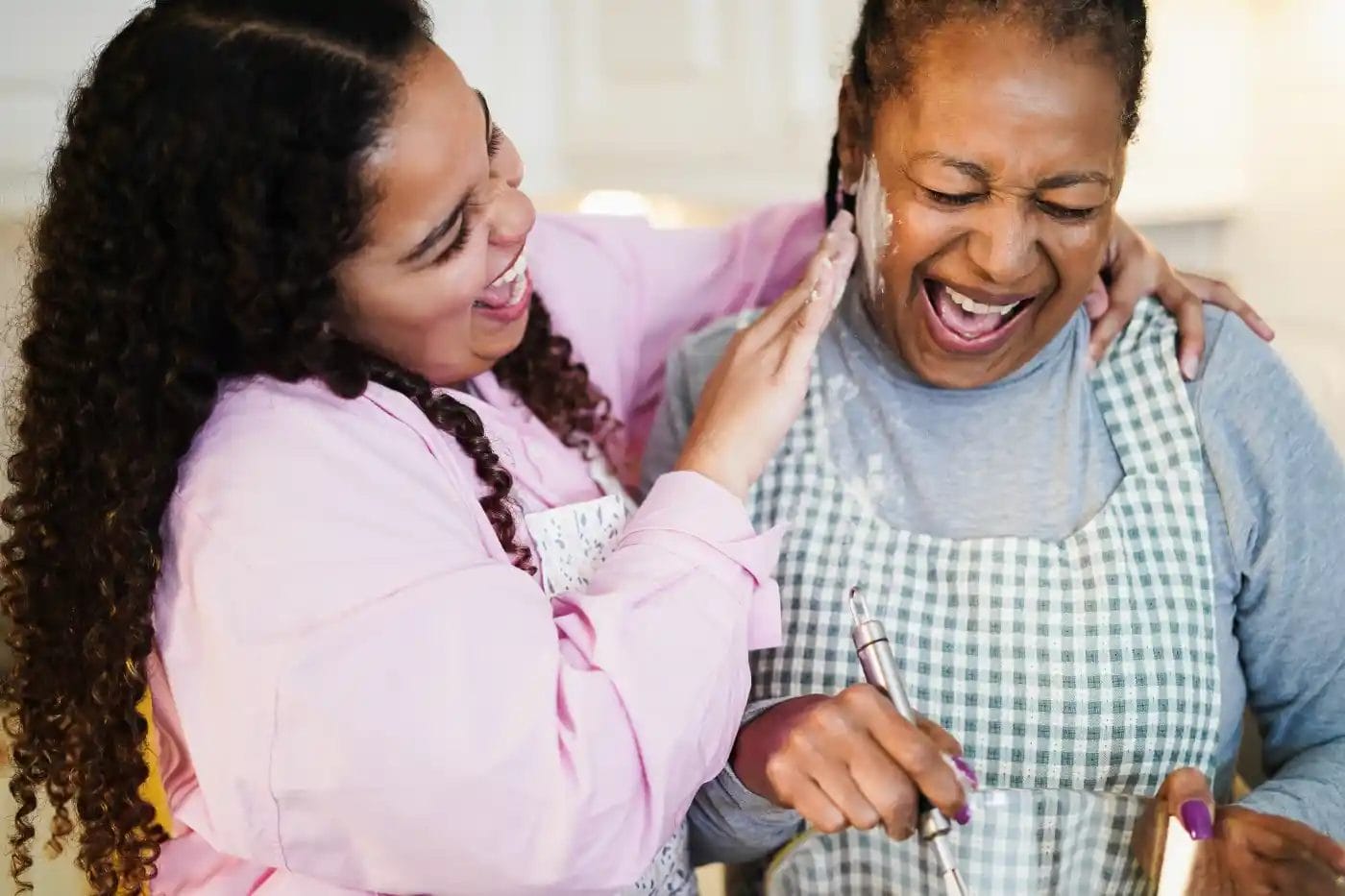 Two women in aprons laugh together in a kitchen while one playfully wipes flour from the other's face.