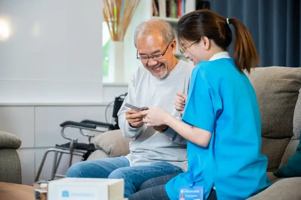 An elderly man sits on a couch smiling and looking at a card, while a healthcare worker in blue uniform—providing home care services—sits beside him. A wheelchair is visible in the background.
