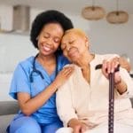 A nurse in blue scrubs sits on a couch beside an elderly woman with a cane, both smiling and leaning their heads together, reflecting the warmth and compassion of quality home care in Columbus.