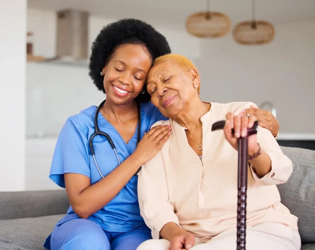 A healthcare worker in scrubs sits beside an elderly woman with a cane, both smiling and showing affection on a couch, capturing the compassionate spirit of Home Care Columbus in a warm home setting.
