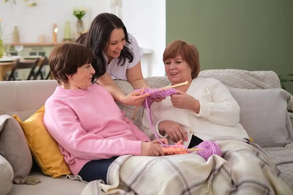 Two older women sit on a couch knitting with chunky yarn while a younger woman from home care services leans over, smiling and observing their work.