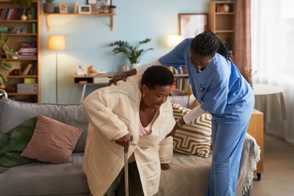 A healthcare worker provides in home care in Columbus, Ohio, assisting an older woman with a cane as she stands up from a couch in a well-lit living room.