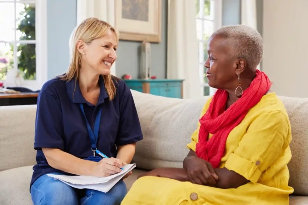 A woman in a navy shirt holding a clipboard smiles while discussing programs with another woman in a yellow outfit and red scarf, as they sit together on a sofa in a bright room.