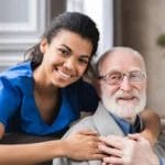A young woman in blue scrubs smiles and embraces an older man with glasses and a beard as they sit together indoors, reflecting the compassionate spirit of Respite Care in Columbus Ohio.