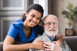 A young woman in blue scrubs smiles and embraces an older man with glasses and a beard as they sit together indoors, reflecting the compassionate spirit of Respite Care in Columbus Ohio.