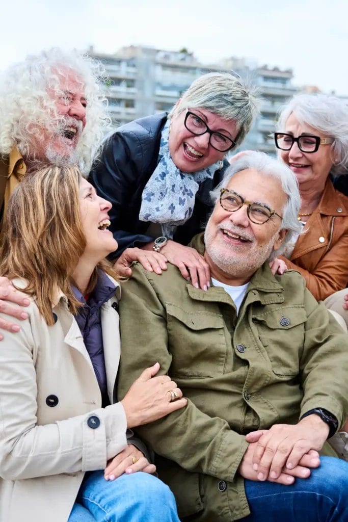 Five older adults, both men and women, wearing casual jackets and glasses, sit closely together outdoors, smiling and laughing—enjoying the warmth of companionship that quality Home Care Columbus can provide.