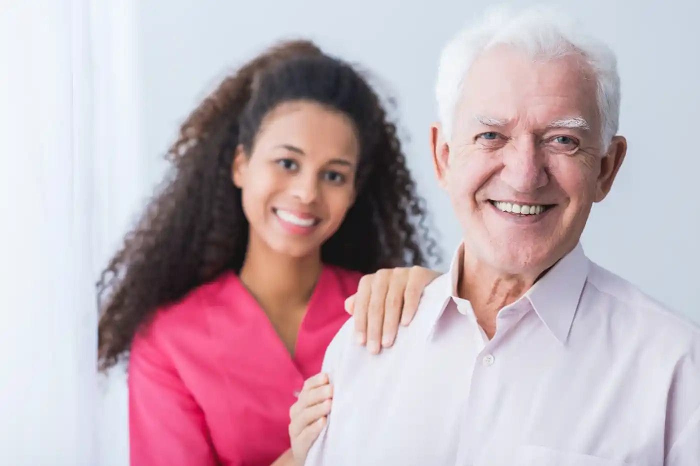 Smiling older man in a light shirt stands in front of a young woman in pink scrubs, who rests her hand on his shoulder. Both are looking at the camera, reflecting the warmth and support of quality home care services.
