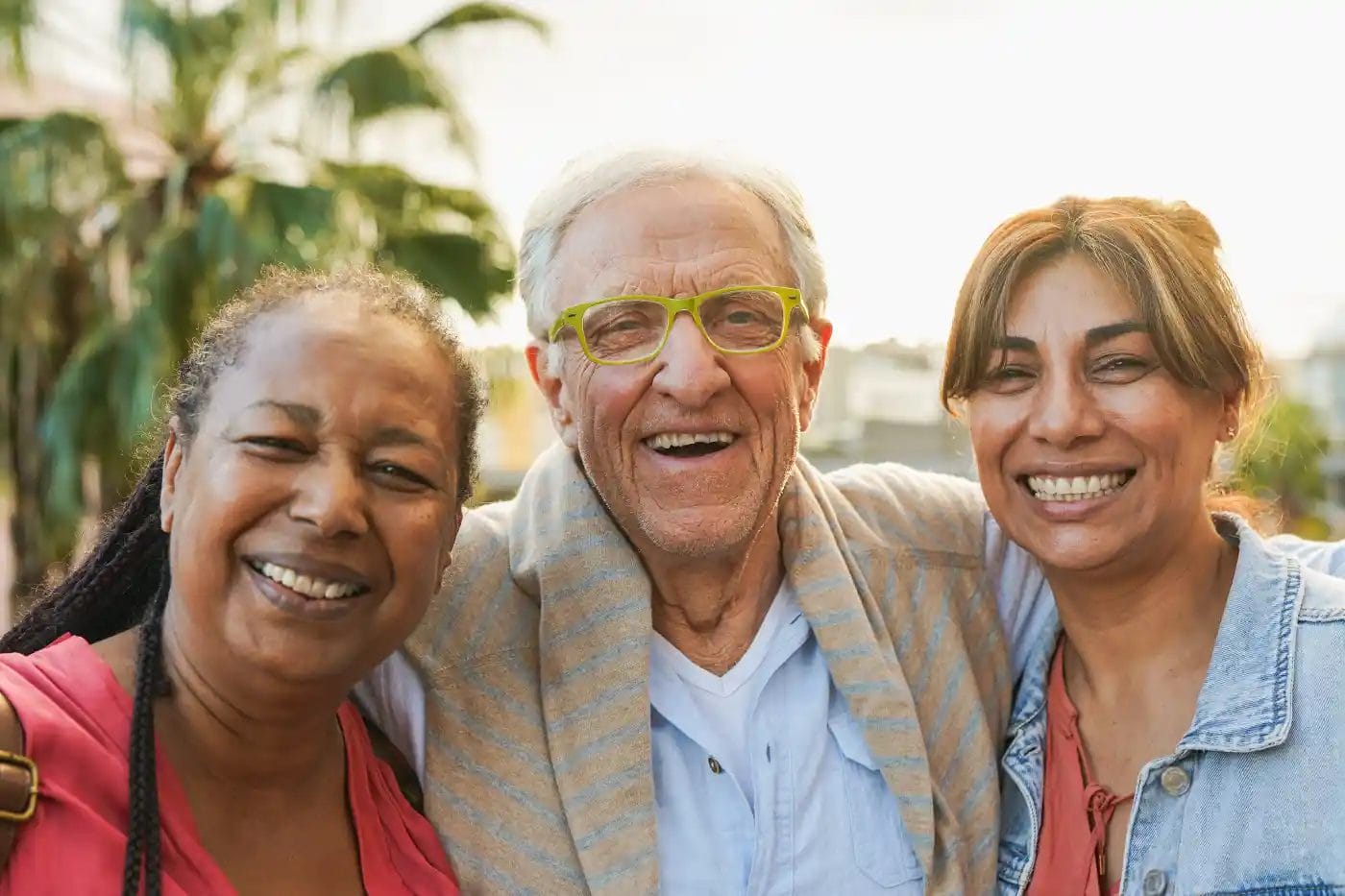 Three adults stand close together outdoors, smiling at the camera. With palm trees behind them and warm late afternoon light, this friendly scene captures the spirit of “Why Us”—genuine connection in a welcoming environment.