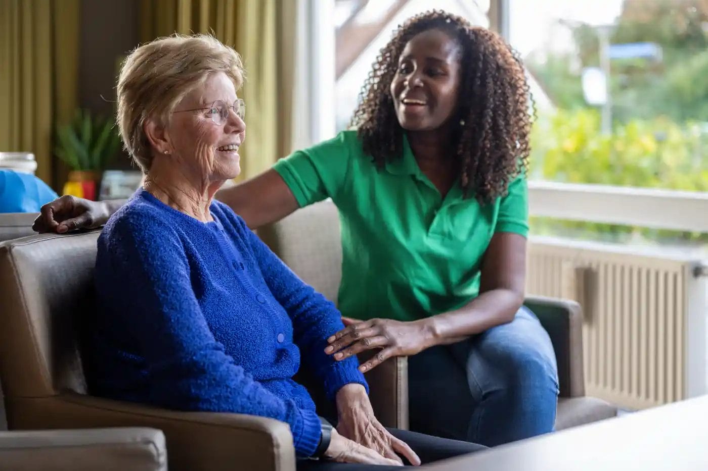 An older woman sits on a chair smiling while a younger woman in a green shirt, visiting for post hospital care, sits beside her talking and smiling in a bright living room.