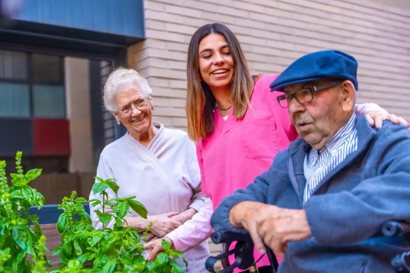 A young caregiver stands with two elderly people by a garden, smiling together outdoors at an assisted living Columbus Ohio community. The elderly man sits with a walker, while the woman stands beside him.