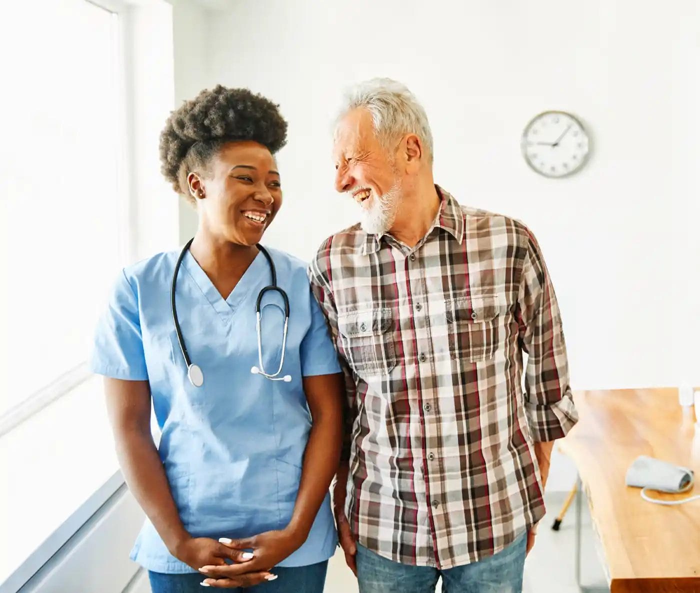 A nurse in blue scrubs stands next to an older man in a plaid shirt, both smiling in a bright room with a wall clock—capturing the warmth of Non Medical Home Care.