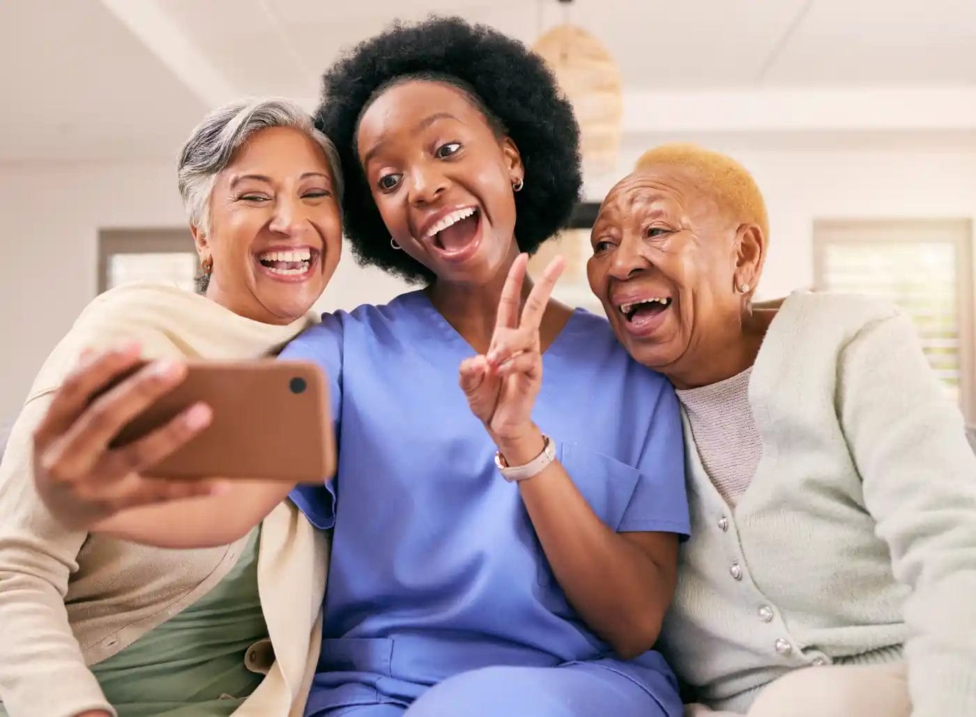 Three women, one in scrubs, sit closely together on a couch, smiling and taking a selfie; one woman flashes a peace sign.