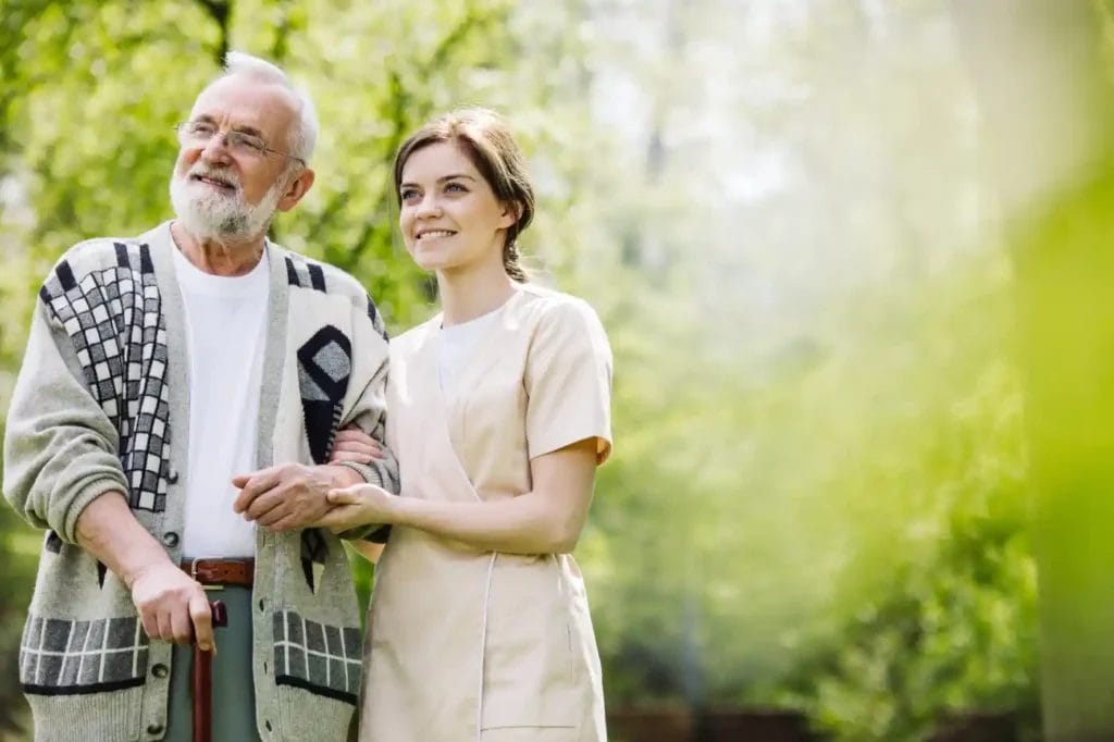 An elderly man with a cane walks outdoors with a young female caregiver in a light uniform. They are smiling and standing among greenery, enjoying companionship and support alongside reliable medication management services.