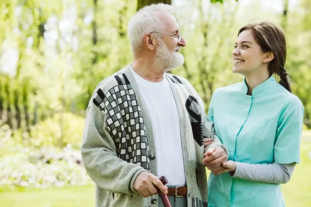 An elderly man with a walking stick and a young woman in a light blue uniform, sharing a smile outdoors, illustrate how compassionate resources can support joyful moments together.