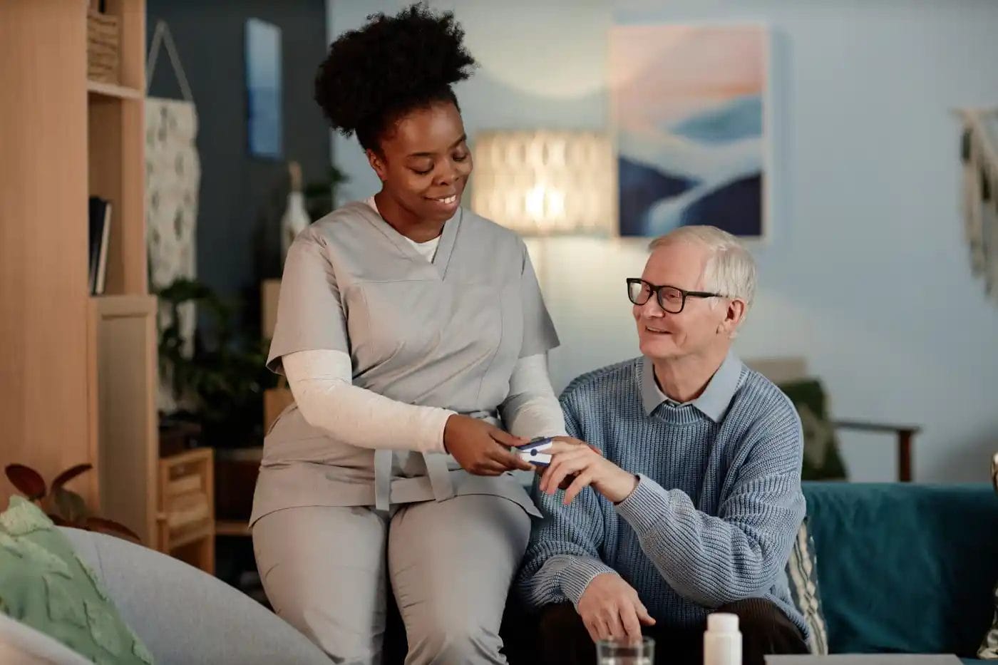 A caregiver in scrubs assists an older man with medication in a living room setting.
