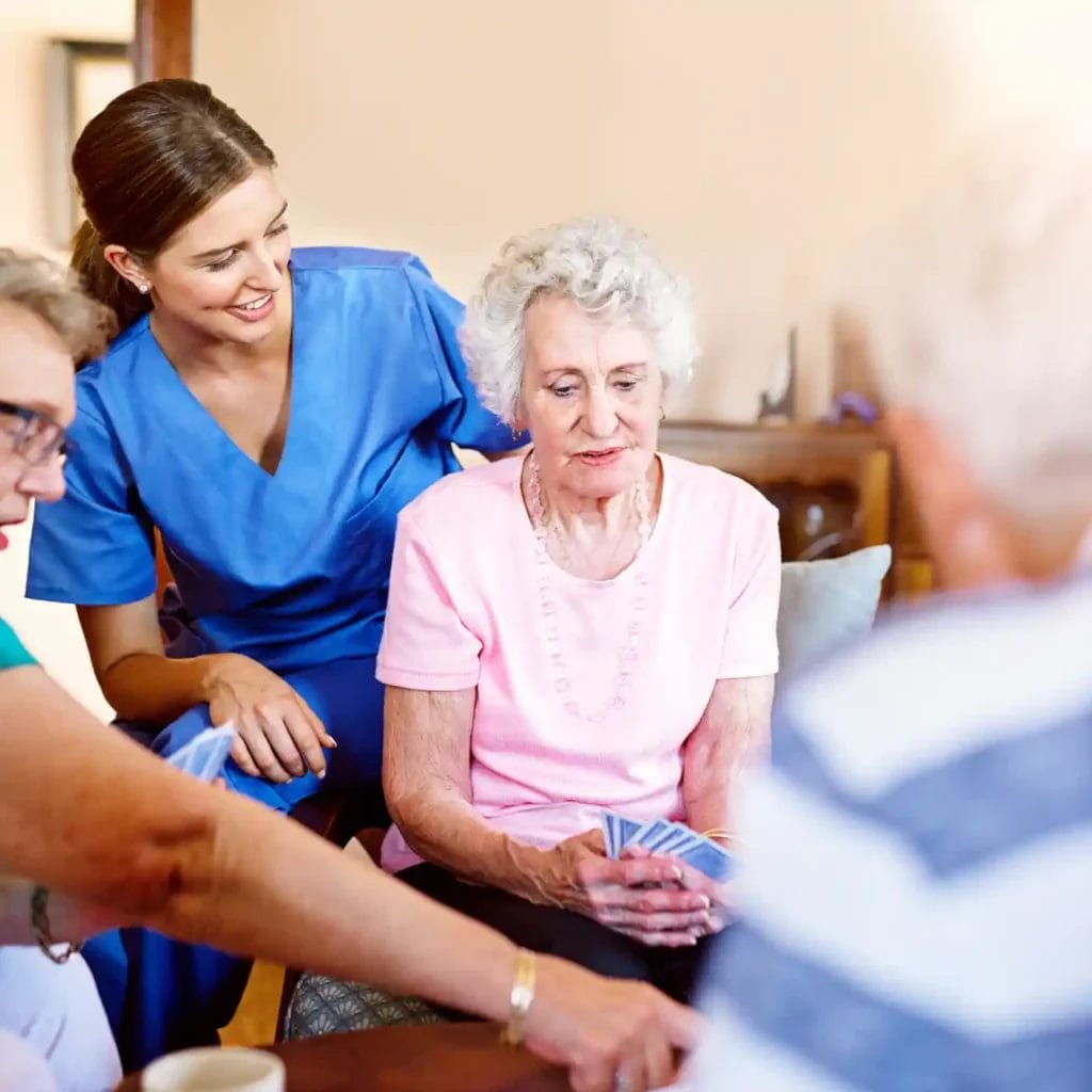 A nurse in blue scrubs, providing home care services, assists elderly people playing cards around a table in a well-lit room.