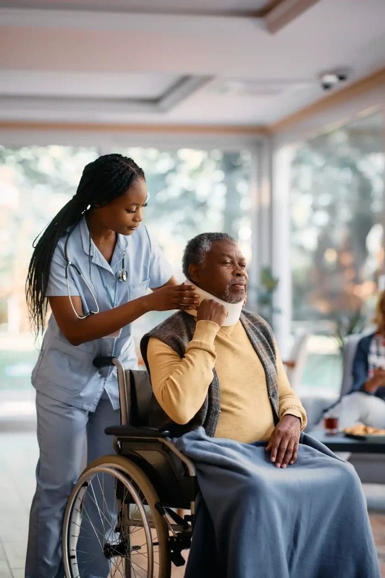 A healthcare worker adjusts a neck brace on an older adult sitting in a wheelchair, covered with a blanket, in a well-lit room where medication management services are also provided.